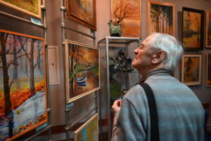 Starszy mężczyzna przygląda się obrazom wywieszonym na ścianie. Jego wzrok jest skupiony. Wydaje się podziwiać jesienny krajobraz, który mieści się na wysokości jego wzroku. 