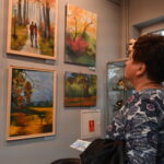 Dorosła kobieta stoi przed ścianą z obrazami przedstawiającymi jesienny krajobraz. Jej wzrok jest skupiony na jednym z obrazów, na którym przedstawiona jest para przechadzająca się aleją drzew w parku.