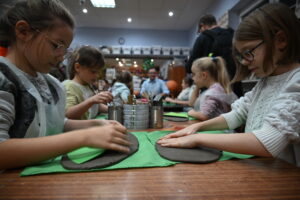 Grupa dzieci siedzi przy stolikach na przeciwko siebie. Dzieci mają przed sobą okrągłe placki z gliny. Dłońmi nadają im kształty. Na twarzach dzieci widać zadowolenie i skupienie.
