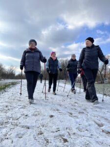 Grupa dorosłych kobiet idzie przed siebie. W rękach trzymają kije do Nordic Walking i spacerują. Na ich twarzach widać uśmiech, kobiety rozmawiają ze sobą wędrując ścieżką po śniegu.