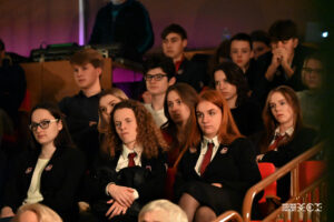 Grupa młodych ludzi w mundurkach szkolnych. Siedzą na fotelach na rozstawionej widowni. Są zasłuchani.