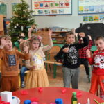 Grupa małych dzieci stoi jedno obok drugiego w klasie szkolnej, przy okrągłym stoliku. Dzieci podnoszą swoją prawą rękę ku górze, jakby zgłaszały się do odpowiedzi.