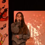 Młoda dziewczyna stoi na kolorowo ozdobionej scenie. W ręku trzyma mikrofon na wysokości ust, wykonuje gest przypominający śpiewanie.