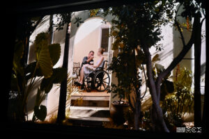 Na pierwszym planie drzewa i wysokie ogrodowe rośliny. W głębi taras przed domem. W centrum kobieta i mężczyzna. Kobieta siedzi na wózku inwalidzkim, mężczyna podaje jej jedzenie.