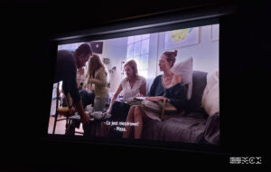 Ekran kinowy. Na nim kadr z filmu przedstawiający trzy kobiety siedzące na kanapie. Przed nimi obok stolika stoi mężczyzna. Podaje im coś do picia.