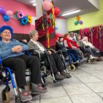 Grupa seniorek na wózkach inwalidzkich stoi w rzędzie obok siebie. Wzrok kobiet skierowany jest na wprost. Na ich twarzach widać uśmiechy i zadowolenie.
