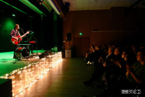 Duża sala ze secną i widownią. światło jest przygaszone, scena ozdobiona świecącymi się lampkami. Na scenie stoi mężczyzna z gitarą. Na widowni siedzą ludzie.