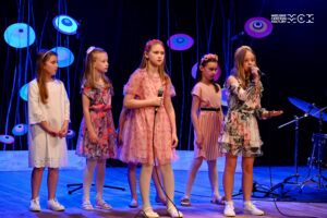 Kilka dziewczynek w wieku szkolnym stoi na scenie. Dwie z nich trzymają w rękach mikrofony. Jedna do niego śpiewa.