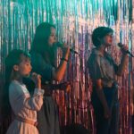 Trzy młode dziewczyny stoją na kolorowo ozdobionej scenie obok siebie. Każda z nich w rękach trzyma mikrofon, do którego zdaje się śpiewać. Wzrok kierują przed siebie, a na ich twarzach widać skupienie.