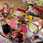 Grupa dzieciaków siedzi przy kolorowym stoliku, na którym rozłożone są kredki oraz kolorowanki. Dzieci jednak spoglądają w kierunku aparatu, machając rękoma i szeroko uśmiechając się. Na ich twarzach widać dobrą zabawę.