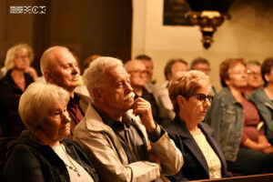 Kilka siedzących w ławach kościelnych osób. Są skupieni, zasłuchani.