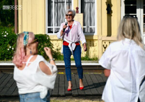 Kobieta w wieku senioralnym stoi na tle jakiegoś budynku. W ręku ma mikrofon, do którego śpiewa lub mówi. Jest elegancko ubrana. Przed nią stoi publiczność.