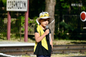 Dziewczyna w kapeluszu. Stoi na tle tablicy z napisem "Czarnocin". W ręku trzyma mikrofon, do którego śpiewa lub mówi. Patrzy przed siebie, jest skupiona.
