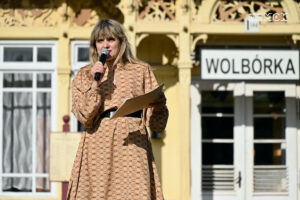 Młoda kobieta stoi na tle zabytkowego drewnianego budynku. świeci słońce. W jednej ręce kobieta trzyma mikrofon, do którego coś mówi, w drugiej kartkę papieru.