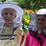 Młody chłopiec z dziewczynką pozują do zdjęcia. Na sobie mają pszczelarskie kapelusze. Na ich twarzach widać szerokie uśmiechy i zniecierpliwienie.