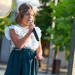 Mała dziewczynka stoi na scenie w przestrzeni miejskiej. W ręku trzyma mikrofon, do którego zdaje się śpiewać. Na jej twarzy widać mocne skupienie.