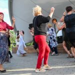 Grupa kobiet w różnym wieku znajduje się w przestrzeni miejskiej. Kobiety zdają się tańczyć, tworząc koło. Ręce trzymają w górze, a ich twarze są zadowolone.
