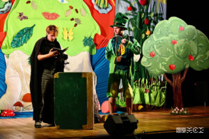 Dwaj mężczyźni stoją na scenie. W tle dekoracja imitująca las. Obaj są przebrani - jeden jest w zielonym stroju przypominającym świerszcza drugi w czarnym. Jest prawdopodobnie pająkeim.