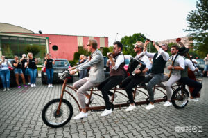 Sześciu mężczyzn siedzi n jednym, sześcioosobowym rowerze. Jeden z nich kieruje, inni mają instrumenty, na których grją podczas jazdy. 