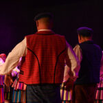 Zespół ludowy wykonuje taniec i przyśpiewki ludowe na kolorowo oświetlonej scenie.