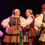 Zespół ludowy wykonuje taniec i przyśpiewki ludowe na kolorowo oświetlonej scenie.