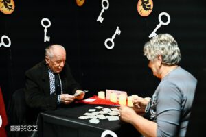 Dwie osoby siedzące naprzeciwko siebie przy kwadratowym stoliku. Mężczyzna trzyma w ręce karty. Na stoliku stoją świece. W tle na ścianie powieszone są wycięte z papieru symbole kluczy.