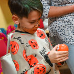 Młody chłopiec w stroju w dynie trzyma w rękach okrągłą kulkę z napisem. Wygląda, jakby skupiał się na przeczytaniu informacji.