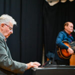 Dwóch starszych mężczyzn znajduje się na scenie. Jeden z nich gra na instrumencie klawiszowym, natomiast drugi gra na gitarze i śpiewa do mikrofonu ustawionego przed nim.