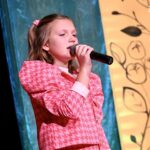 Młoda dziewczynka śpiewa na kolorowo ozdobionej scenie do mikrofonu. Na jej twarzy widać skupienie.