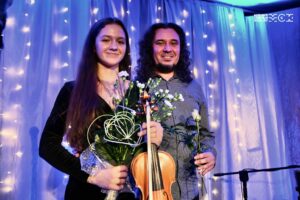 Kobieta i mężczyzna stoją obok siebie na scenie. Kobieta trzyma skrzypce i bukiet kwiatów. Mężczyzna różę. Uśmiechają się.