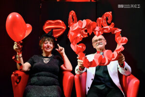 Dwoje seniorów, kobieta i mężczyzna. Kobieta trzyma w ręce czerwony balon w kształcie serca, a mężczyzna ramkę w kształcie serca wyklejona czerwonymi serduszkami. 