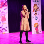 Młoda dziewczynka śpiewa na scenie do publiczności.