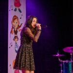 Młoda dziewczyna śpiewa na scenie do publiczności.