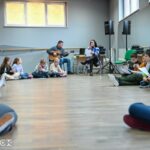 Grupa dzieci siedzi na podłodze w sali lustrzanej. Dzieci siedzą w okręgu wokół instruktorów - młodej kobiety oraz mężczyzny. Wszyscy zdają się śpiewać piosenkę. Dzieci mają przed sobą teksty, kobieta mikrofon, a mężczyzna gra na gitarze.