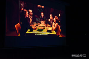 Ekran w kinie z kadrem z filmu, który przedstawia ludzi siedzących przy stole.