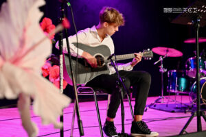 Chłopiec grający na gitarze. Siedzi na krześle na scenie udekorowanej kwiatami.