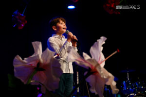 Mały chłopiec śpiewający do mikrofonu. Obok niego dekoracja z kwiatów.