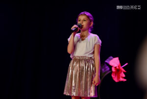 Dziewczynka w w wieku szkolnym stojąca na scenie i śpiewająca do mikrofonu, który trzyma w ręce.