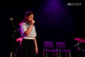 Dziewczynka śpiewająca do mikrofonu, który trzyma w ręce. Stoi na scenie.