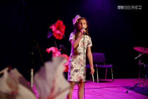 Dziewczynka w w wieku szkolnym stojąca na scenie i śpiewająca do mikrofonu, który trzyma w ręce. Obok niej dekoracja z dużych papierowych kwiatów.