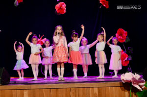 Dzieci na scenie. Jedna dziewczynka, stojąca w środku z mikrofonem, śpiewa. Inne obok niej tańczą.