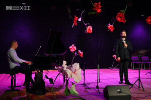 Dwaj mężczyźni na scenie. Jeden gra na fortepianie, drugi śpiewa do mikrofonu stojącego przed nim. Scena udekorowana jest kwiatami.