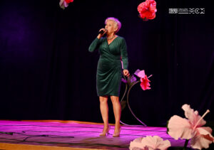 Starsza kobieta na scenie udekorowanej kwiatami. Śpiewa.